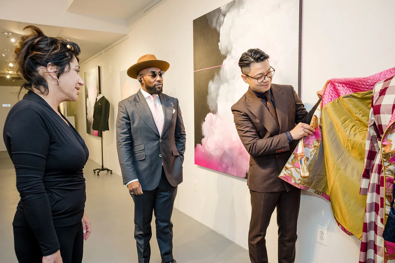 Three people at an art gallery viewing a colorful artwork. One man is unveiling a painting, while a woman and a man observe. The man unveiling is dressed in a brown suit, the woman in black, and the other man in a suit with a hat.