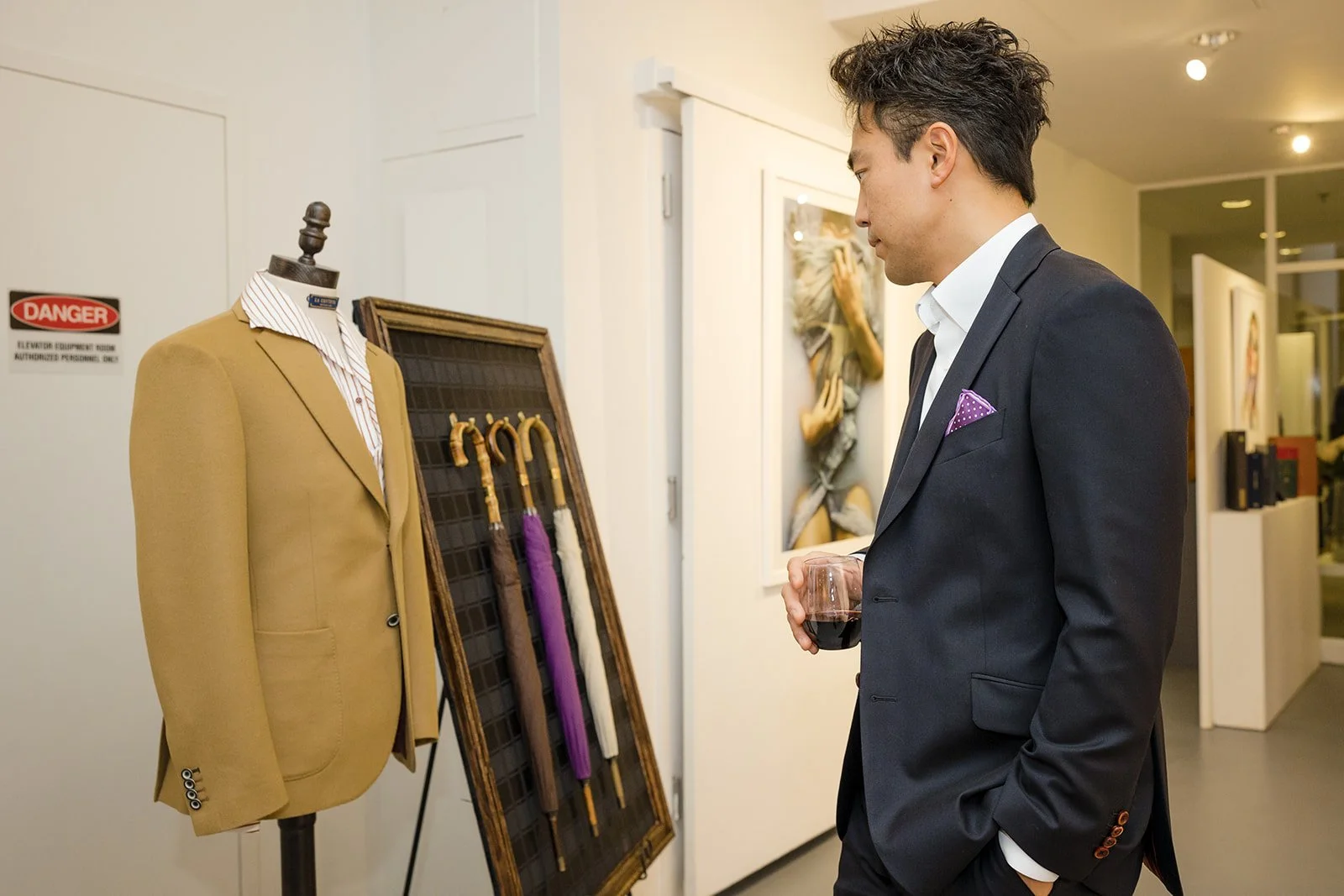 A man in a dark suit holding a glass of red wine, looking at a display of umbrellas and a beige blazer on a mannequin inside an art gallery or boutique store.