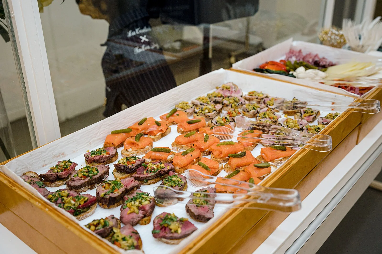 Assorted open-faced appetizers with smoked salmon, steak topped with chopped herbs and vegetables, and other finger foods on a tray with tongs.