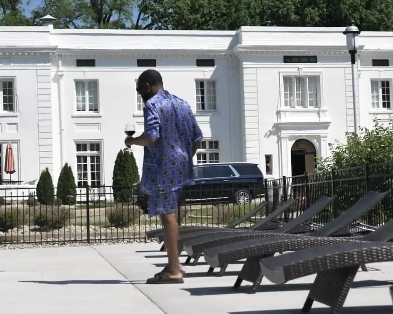 A man wearing a blue patterned shirt and shorts, holding a drink, standing near poolside lounge chairs on a sunny day in front of a white building with large windows.