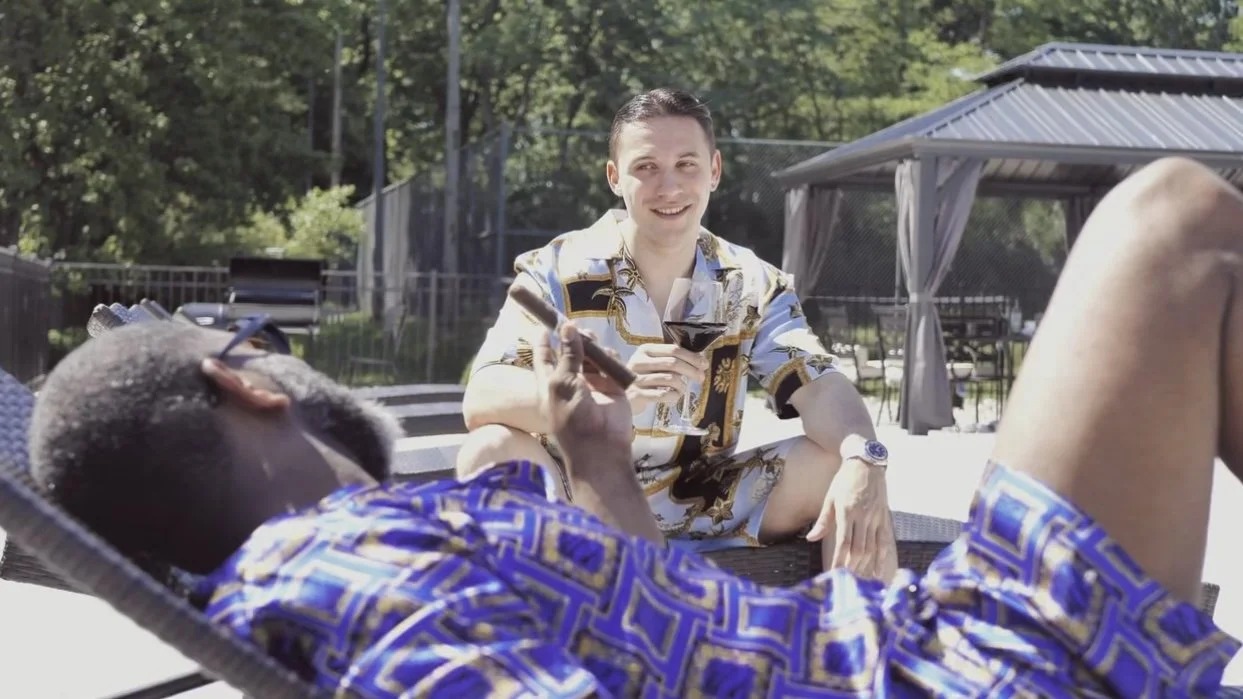 Two men relaxing outdoors on lounge chairs, one laying back and the other sitting and smiling, both wearing colorful summer shirts, in a backyard setting with trees and a fence.