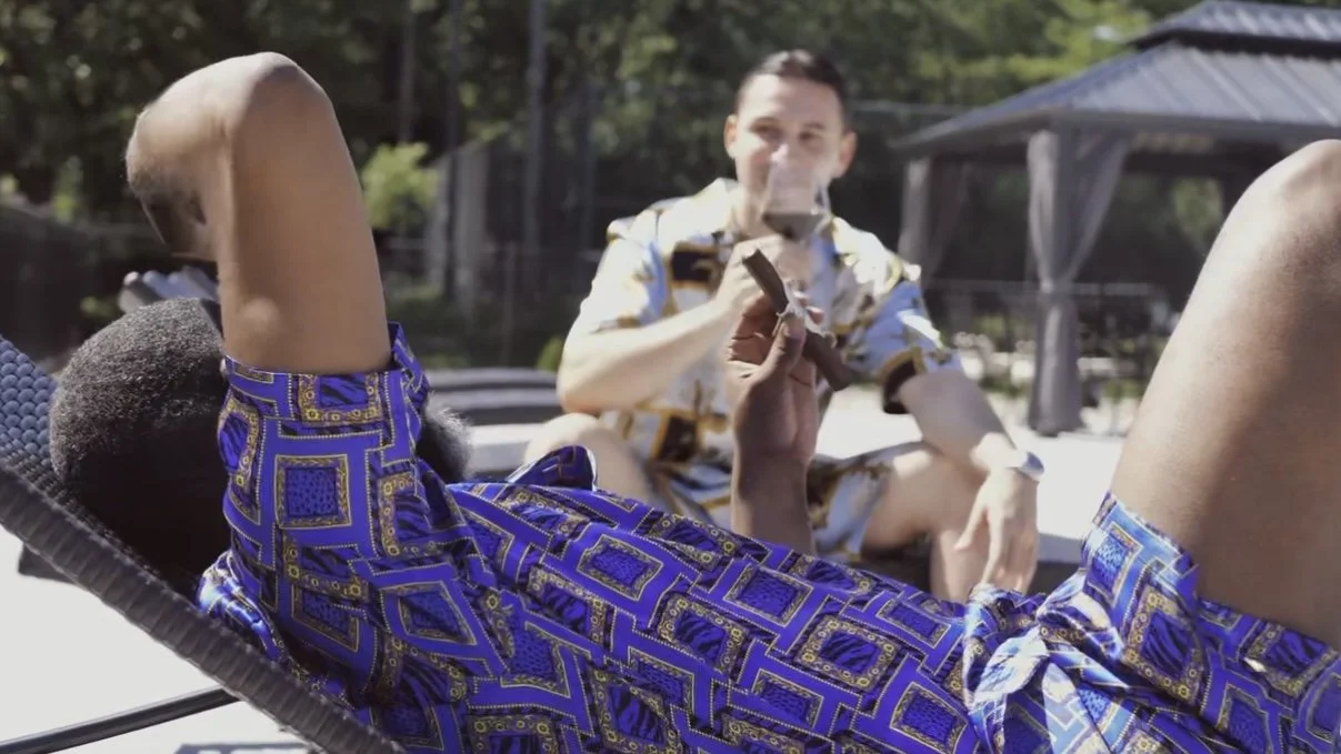 Two men relaxing outdoors, one lying on a lounge chair wearing a colorful patterned shirt and shorts, holding a cigar, and the other sitting, wearing a matching shirt, with trees and a gazebo in the background.