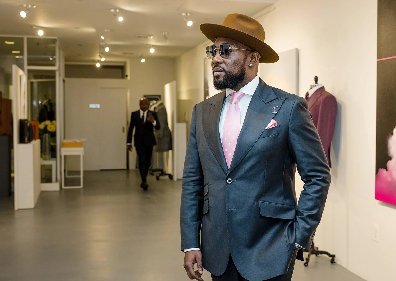 A man wearing a dark suit, pink tie, pink pocket square, sunglasses, and a brown hat, standing inside a modern store or gallery.