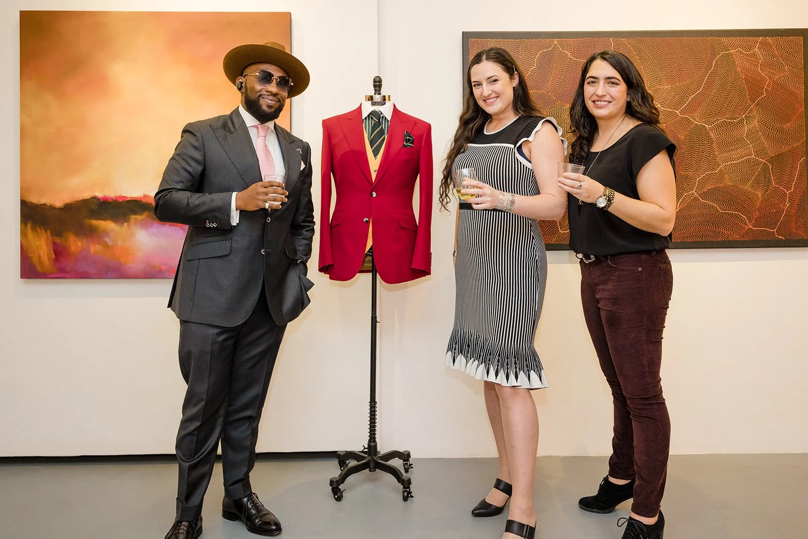 Three people standing in an art gallery, holding drinks, with a suit on a mannequin between them. The man on the left is wearing a dark suit, pink tie, and hat. The woman in the middle is wearing a black and white striped dress. The woman on the righ