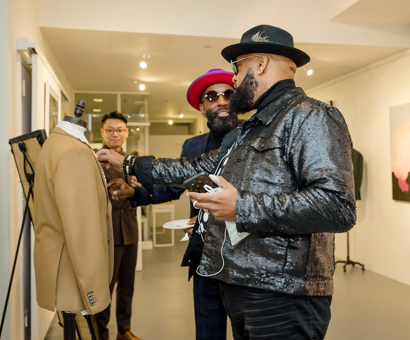 Two men in fashionable clothing looking at a mannequin dressed in a beige blazer and striped shirt, inside an art gallery or boutique, with another person in the background.