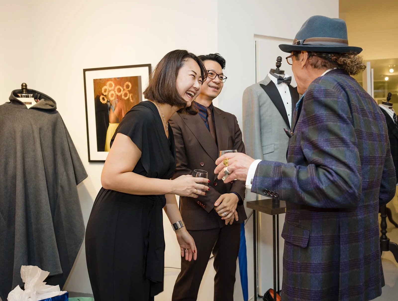 Three people at a social gathering, two women and a man, smiling and holding glasses of wine, engaging in conversation with a man wearing a hat and plaid blazer in a room with clothing displays and framed artwork.