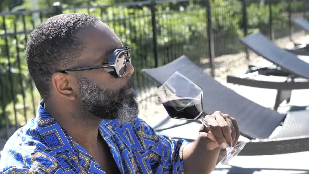 A man with sunglasses, a beard, and a patterned shirt is sitting by a pool holding a glass of red wine.
