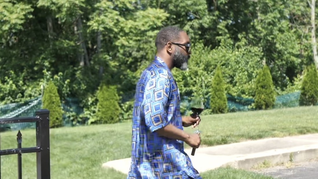 Man walking outdoors in a blue patterned shirt, holding a wine glass with a saucer, in a green backyard with trees and shrubs.