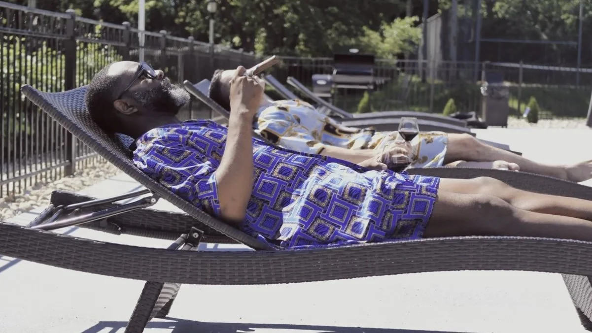Two men are lying on lounge chairs by a pool, one is wearing a patterned blue shirt and the other a floral shirt, both are drinking wine and relaxing.