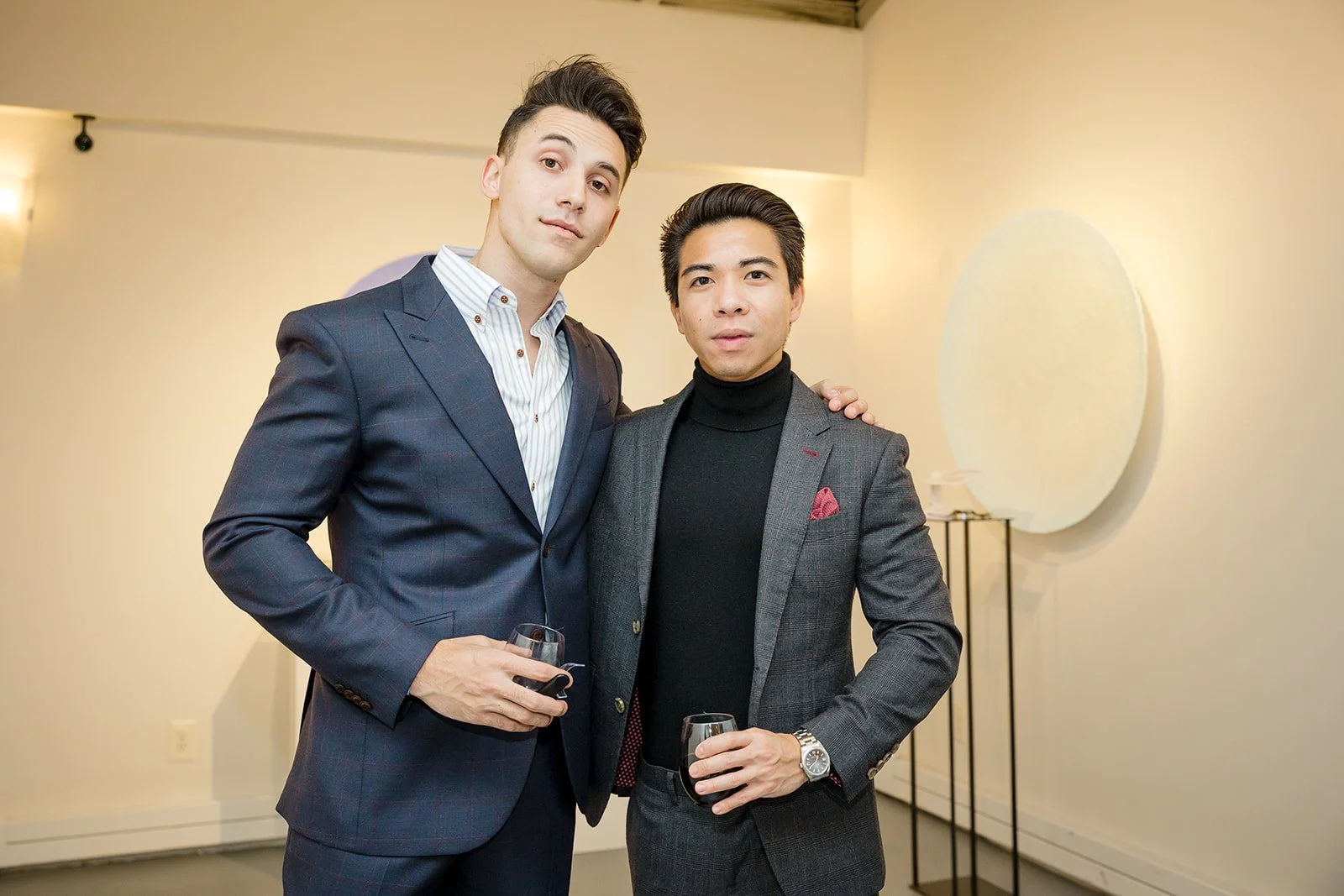 Two men in suits holding glasses of wine at an indoor event.