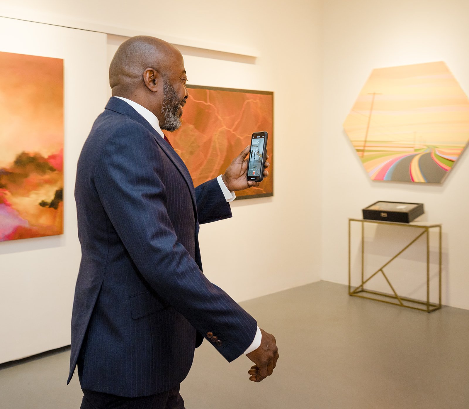 A man in a dark suit takes a photo with his phone inside an art gallery, with abstract landscape paintings on the walls.