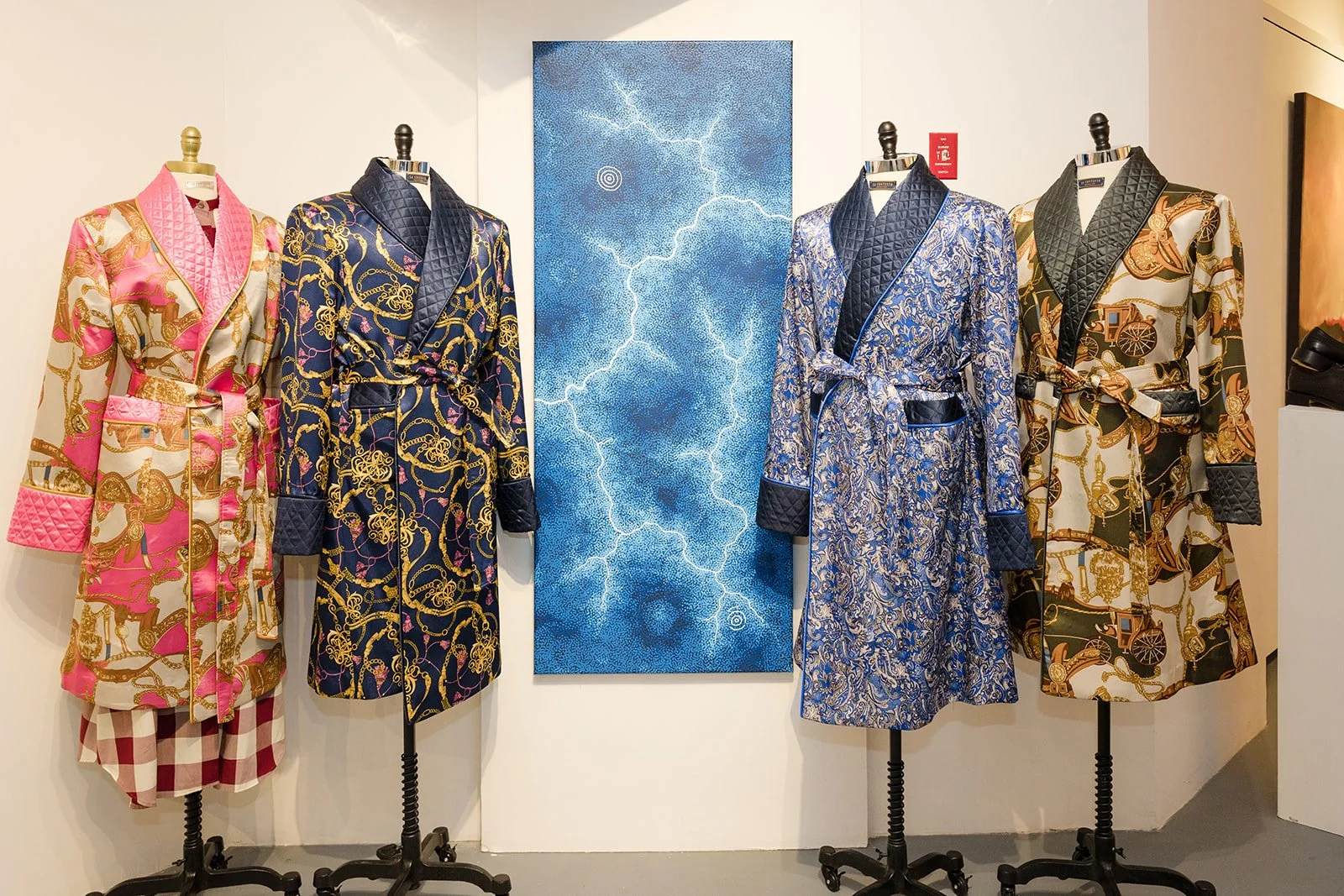 Four mannequins wearing colorful patterned robes with quilted lapels, displayed in front of a blue textured art piece on a white wall.