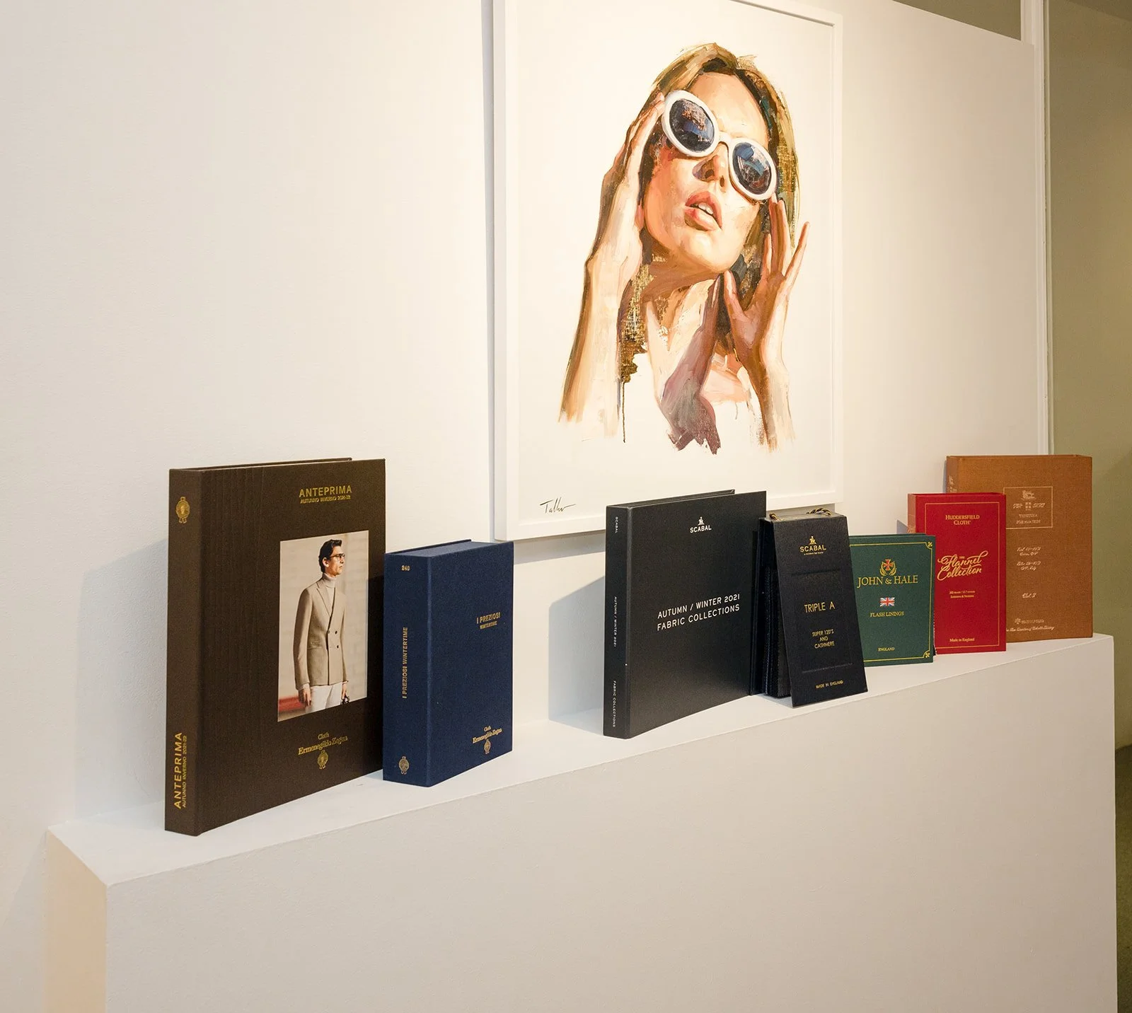 Art print of a woman wearing sunglasses is hung on a white wall above a display of colorful booklets arranged on a white surface.