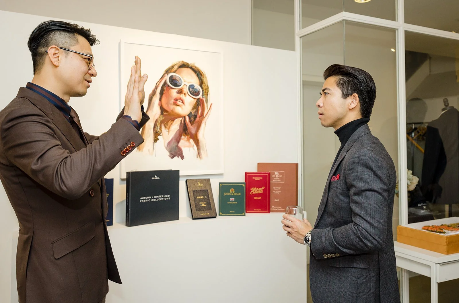 Two men in suits having a conversation at an art gallery, with a portrait of a woman wearing sunglasses hanging on the wall behind them, and several books and folders displayed on a ledge nearby.