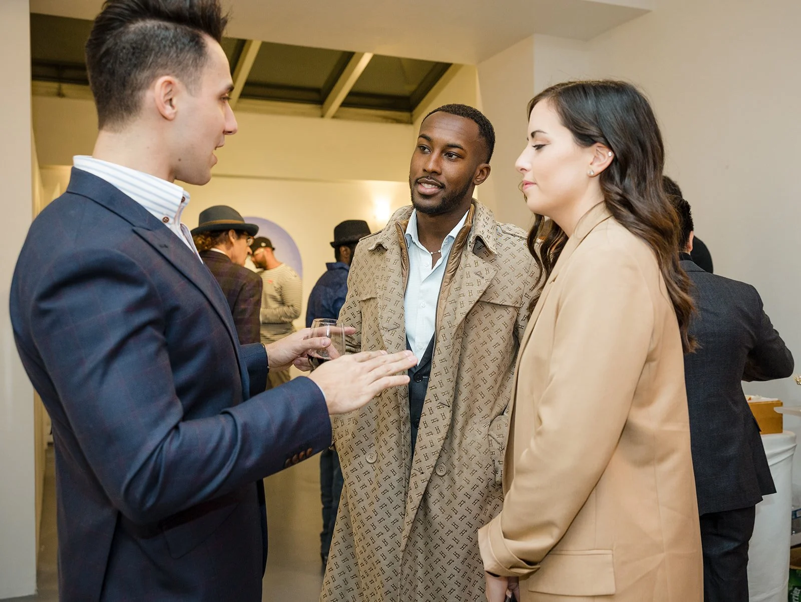 Three people in conversation at a social event, with other guests in the background, indoors.