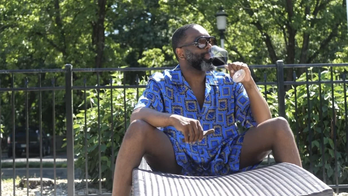 A man in a blue patterned shirt and shorts sitting outdoors on a lounge chair, drinking red wine from a glass, with green trees and a black fence in the background.