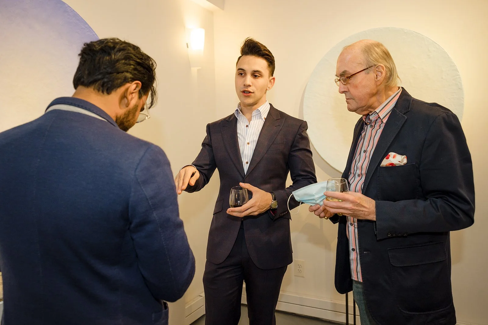 Three men in business attire engaged in conversation at an indoor event. The man on the right is holding a glass of white wine and a face mask, the man in the middle is holding a glass of red wine, and the man on the left has his head bowed, possibly