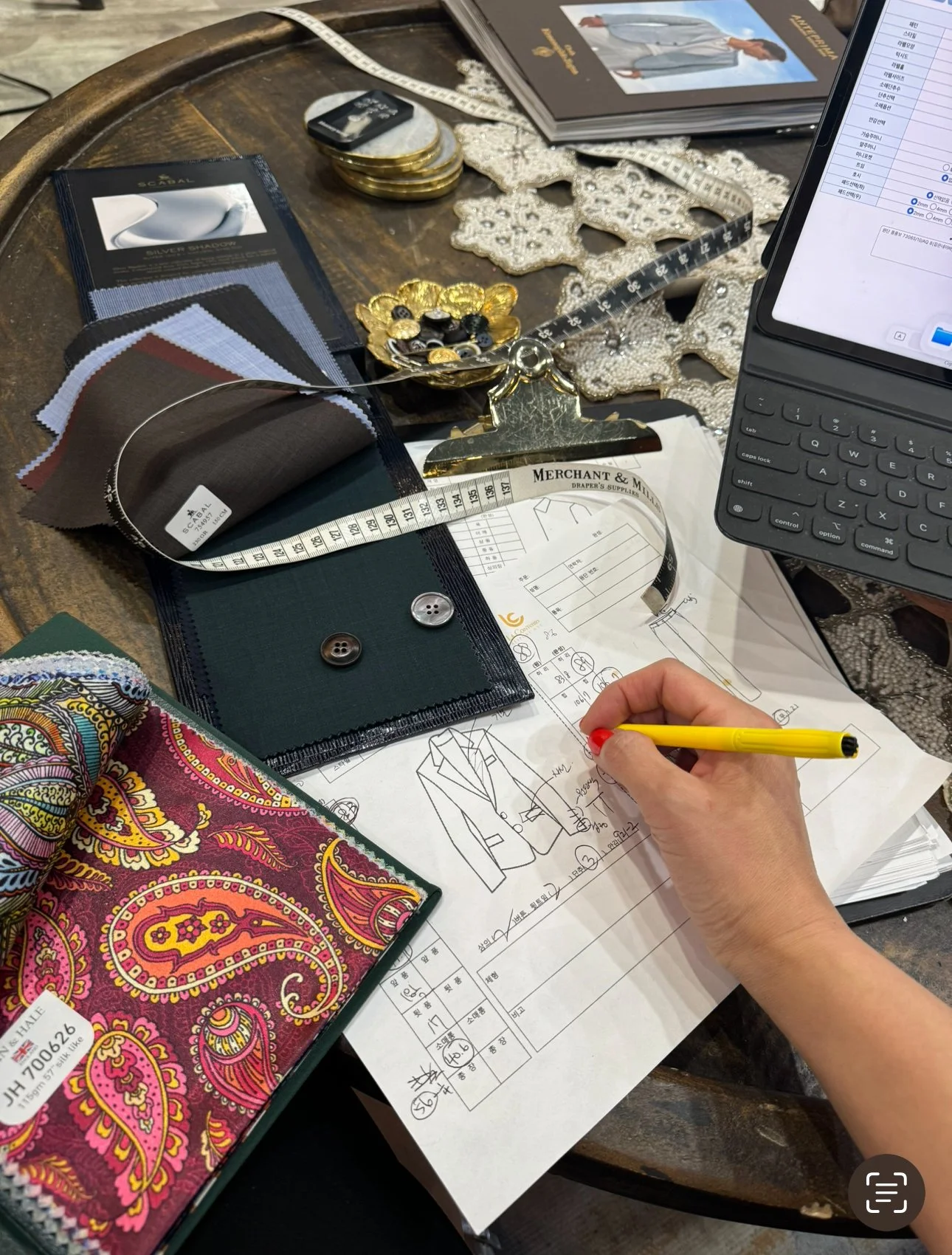 A workspace with fabric swatches, buttons, a tablet with a keyboard, a pattern sketch, and various sewing and design materials.