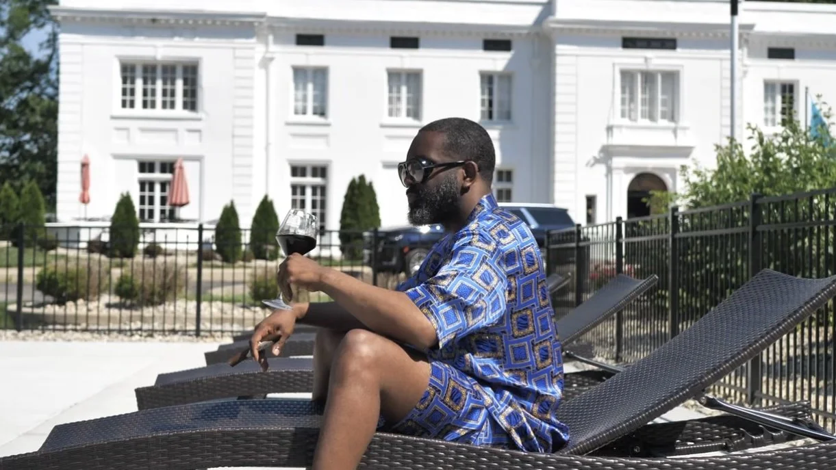 A man with sunglasses and beard relaxing on a lounge chair by a pool, holding a glass of red wine, with a white house in the background.