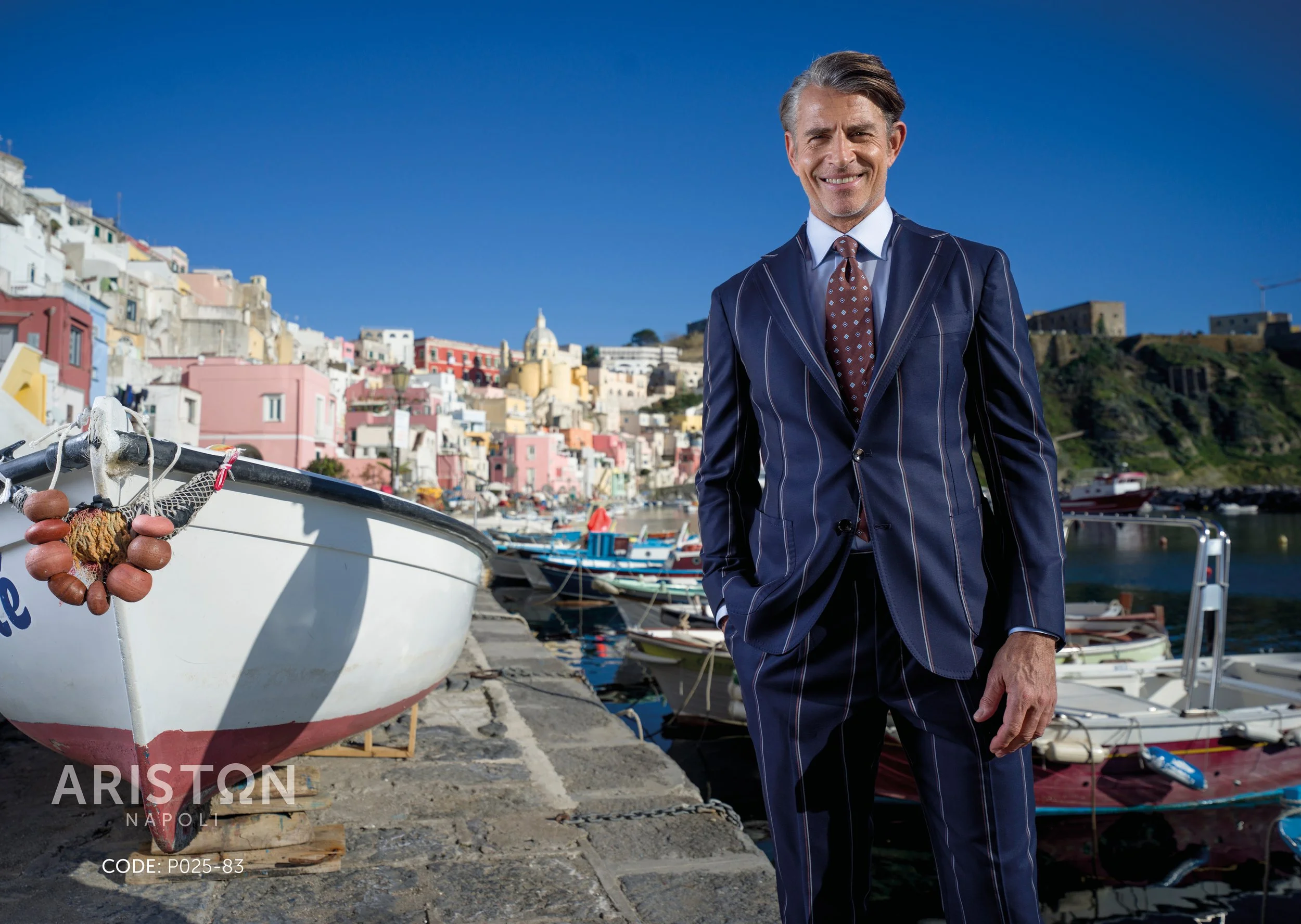 A man in a pinstripe navy suit and patterned tie standing by a marina with colorful houses in the background, under a clear blue sky.