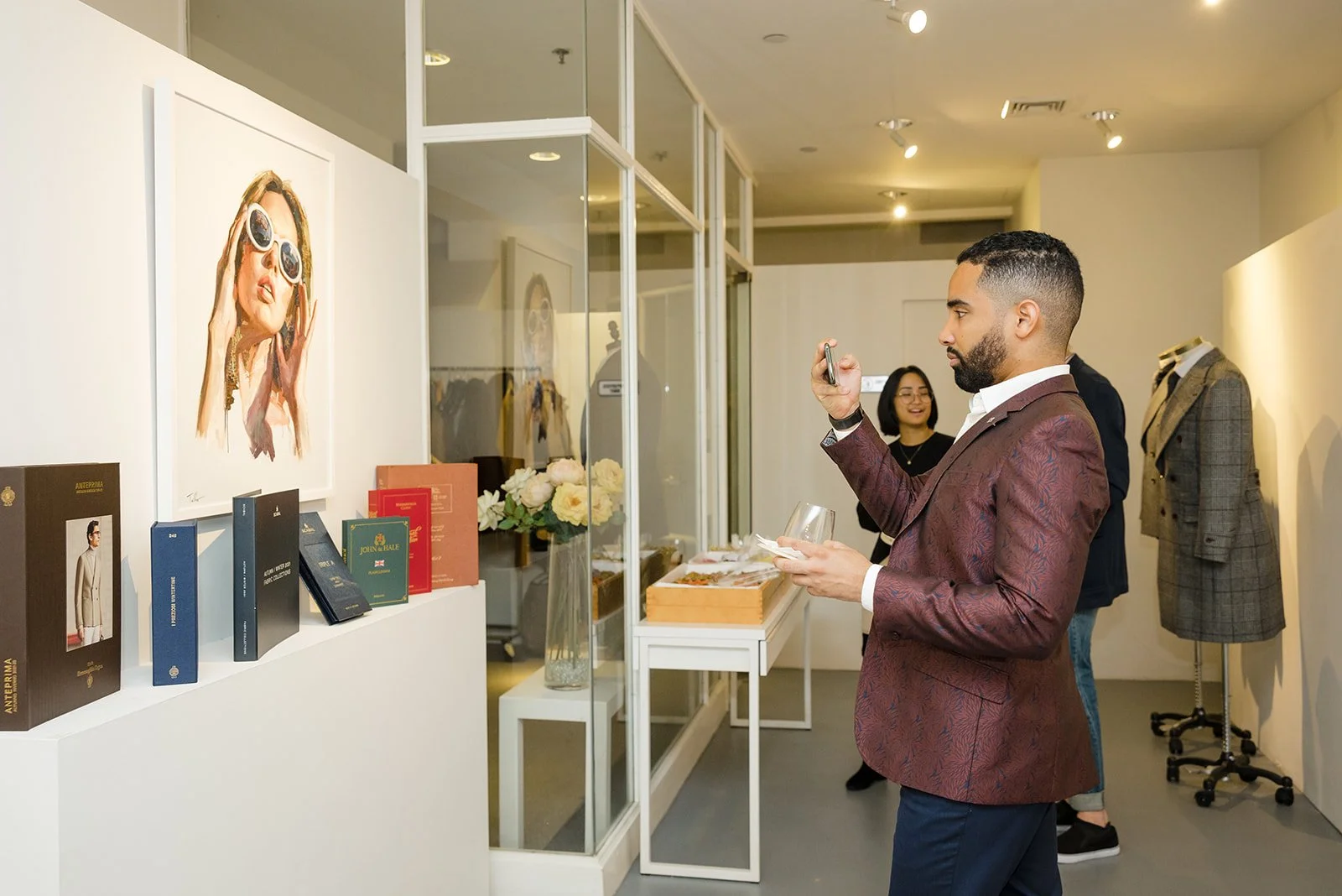 Man in a burgundy blazer taking a photo at an art exhibit with a woman smiling in the background. Art pieces and fashion items on display, including a large portrait of a woman with sunglasses and mannequins with jackets.