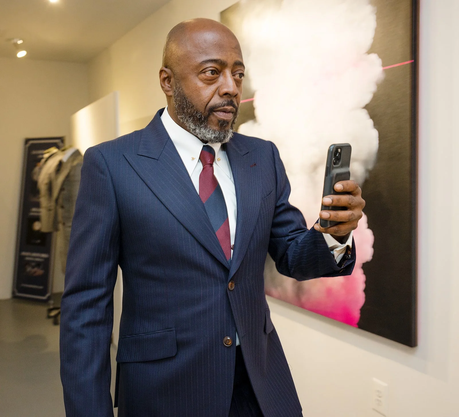 A man in a dark pinstripe suit and tie stands in an art gallery, looking at his smartphone, with a colorful textured artwork behind him.