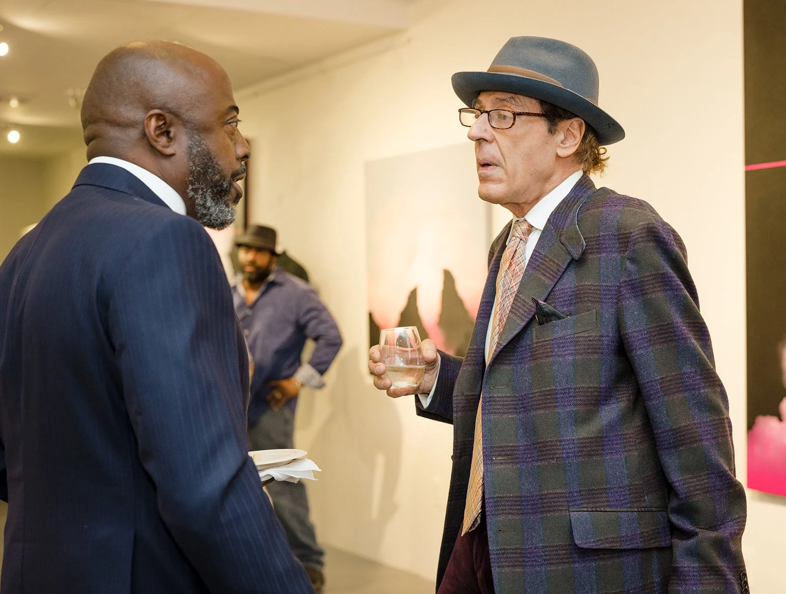 Two men engaged in conversation at an event. The man on the left is dressed in a dark suit with a white shirt and appears to be listening. The man on the right is wearing glasses, a gray fedora hat, a plaid blazer, a shirt, and tie, holding a glass o