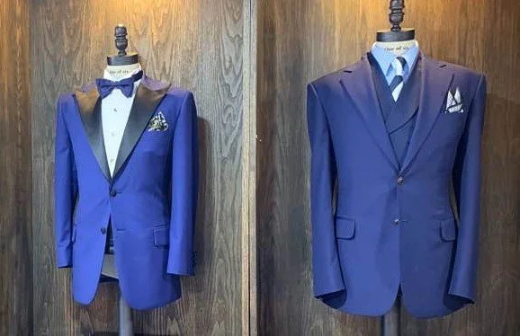 Two blue men's suits on mannequins, one with a bow tie and pocket square, the other with a tie and pocket square, displayed against a wooden background.