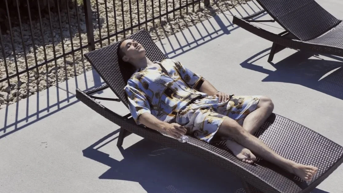 Woman lying on a lounge chair by a fence, wearing a patterned dress, holding a glass, relaxing outdoors in the sun.