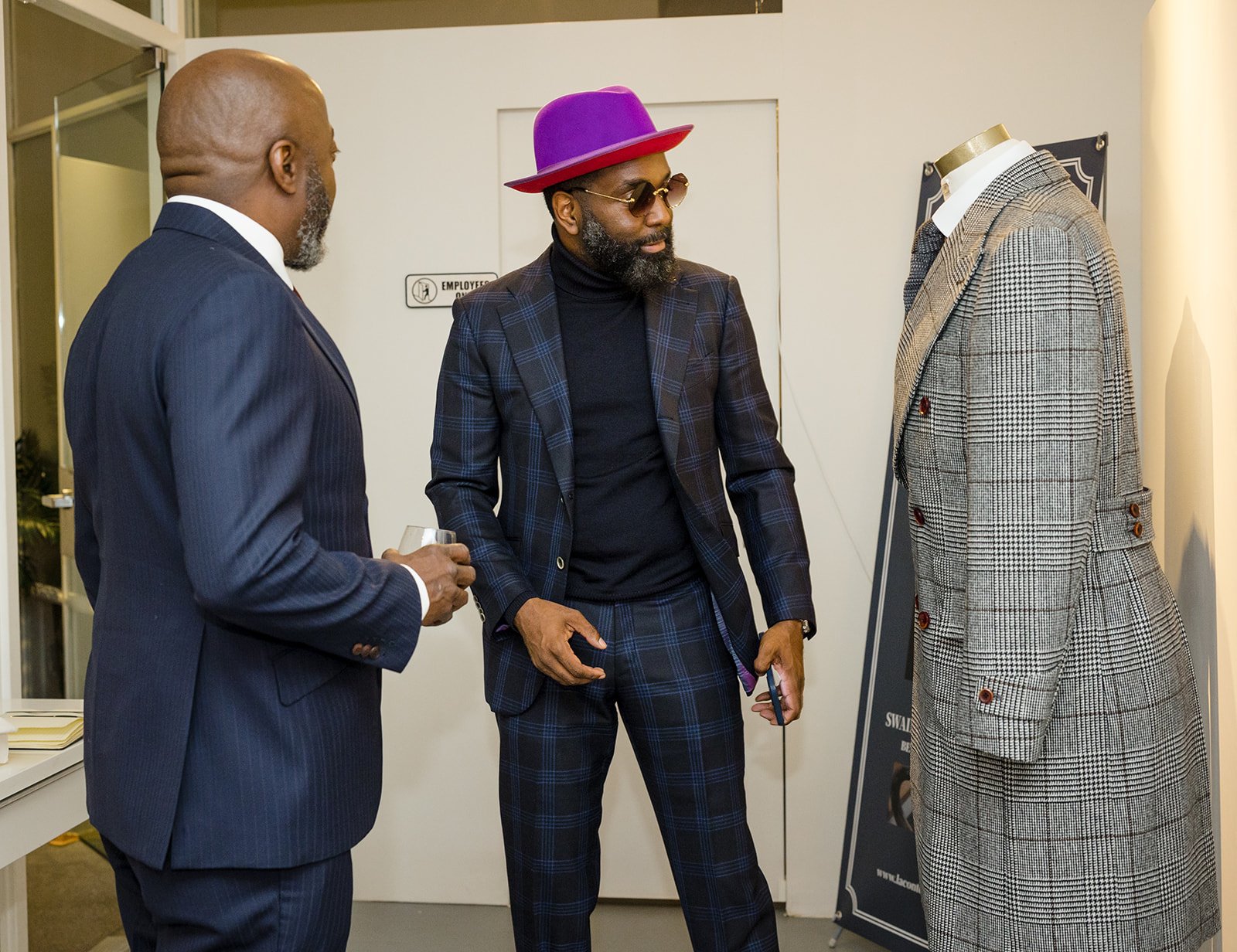 Three men in suits examining a plaid suit on a mannequin in an indoor setting.