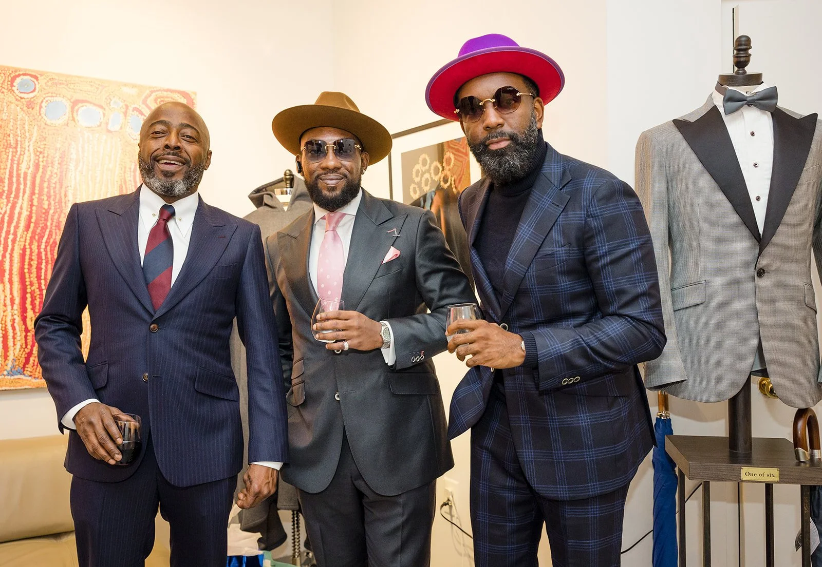 Three men dressed in suits and hats holding drinks at a social event, with mannequins in suits and artworks in the background.