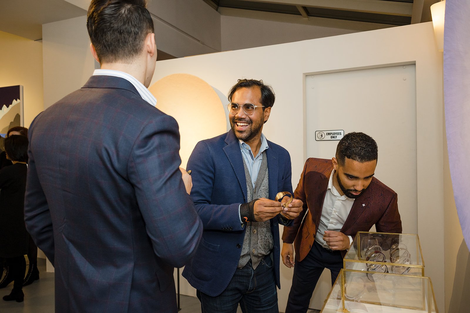 Three men in formal attire talking at an exhibition, with one man smiling and holding a watch, another browsing watches in a glass case, and the third turned away from the camera.