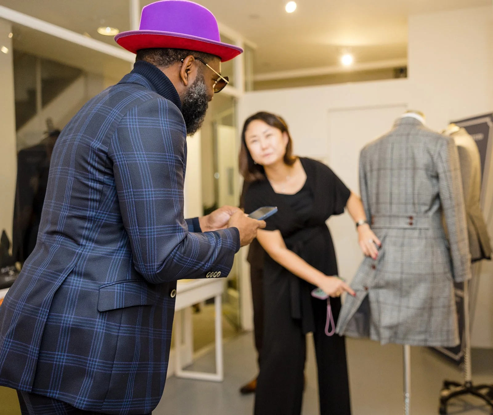A man with a beard wearing a purple and red hat, sunglasses, a dark blue plaid blazer, is looking at his phone in a boutique. A woman in black is pointing at a plaid blazer on a mannequin, which is displayed alongside other blazers.