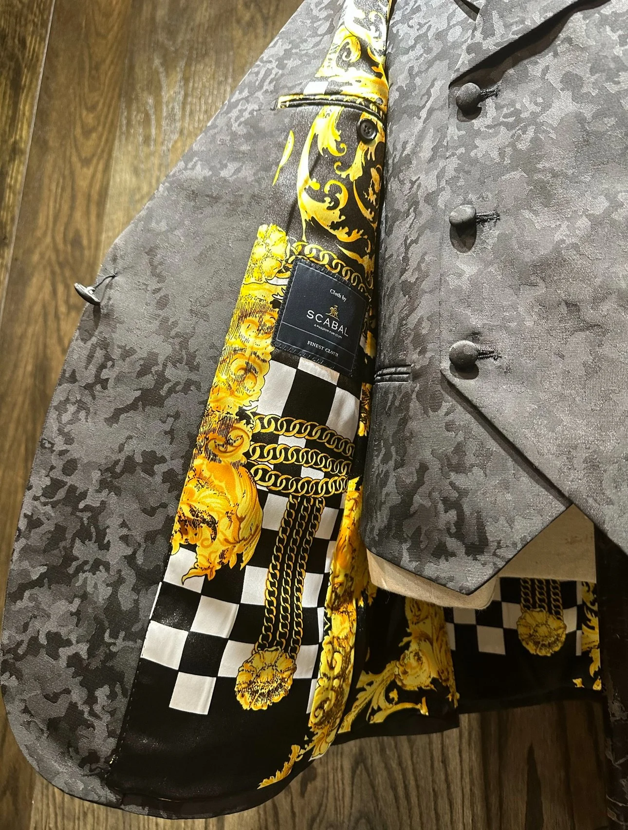 Close-up of layered clothing including a gray patterned blazer, a black and yellow patterned scarf with chain design, and a black label reading 'Cloth by Scabal, Finest Cloth' placed on a wooden surface.