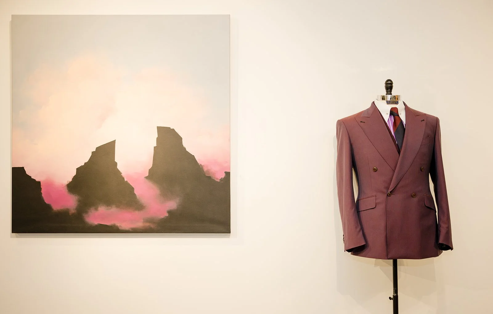 An art gallery display with a painting of a mountain landscape and a vintage mauve suit on a mannequin.