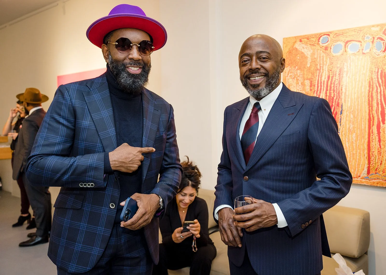 Two men in suits smiling at a social event, one pointing at the other, with a woman in the background looking at her phone and a colorful abstract artwork on the wall.