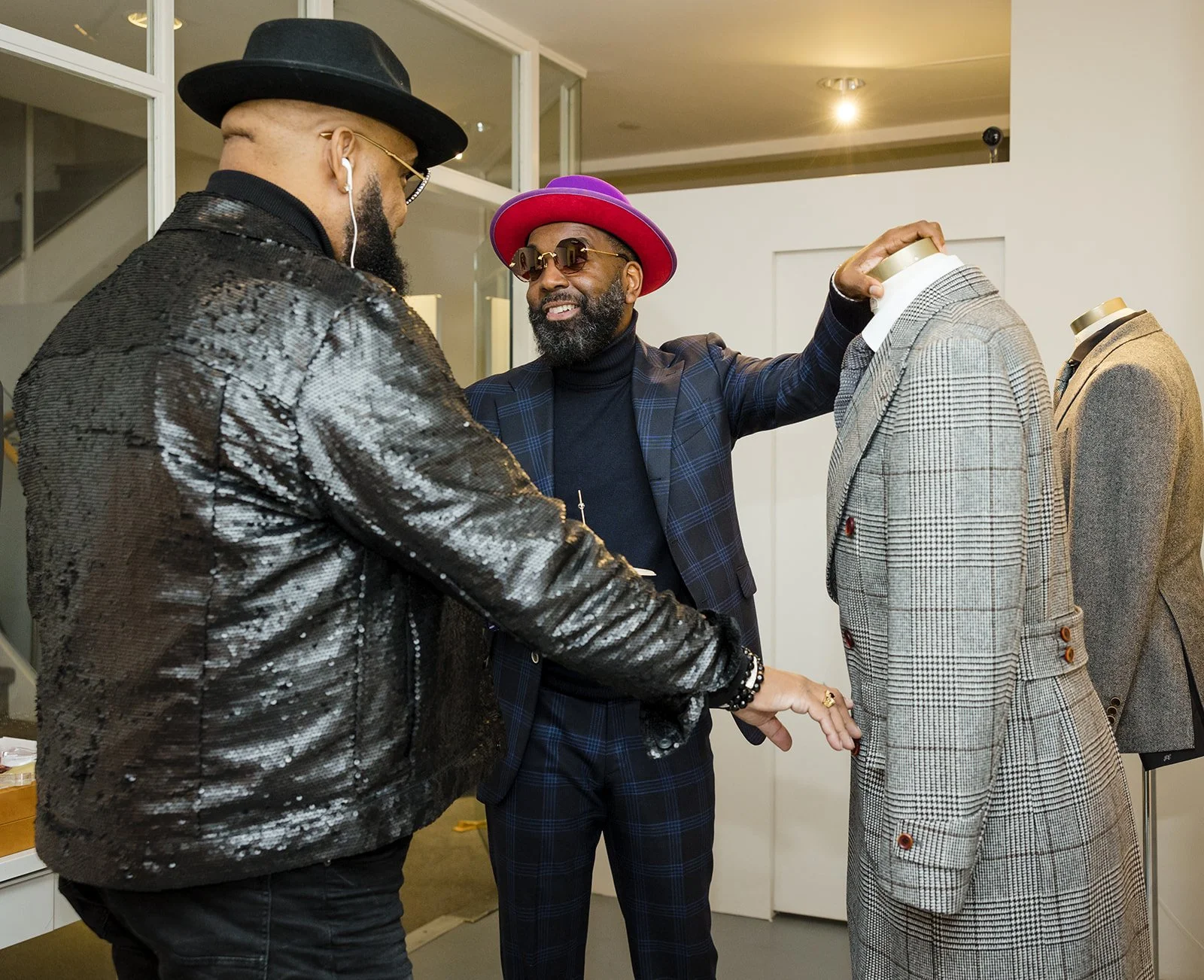 Three men in fashionable clothing, one touching a mannequin dressed in a plaid suit, another wearing a purple hat and sunglasses, and the third in a black shiny jacket, all engaged in conversation in a modern room.
