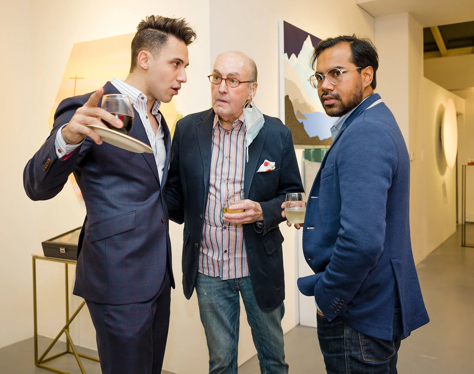 Three men in suits having a conversation, holding drinks at an art gallery or museum.