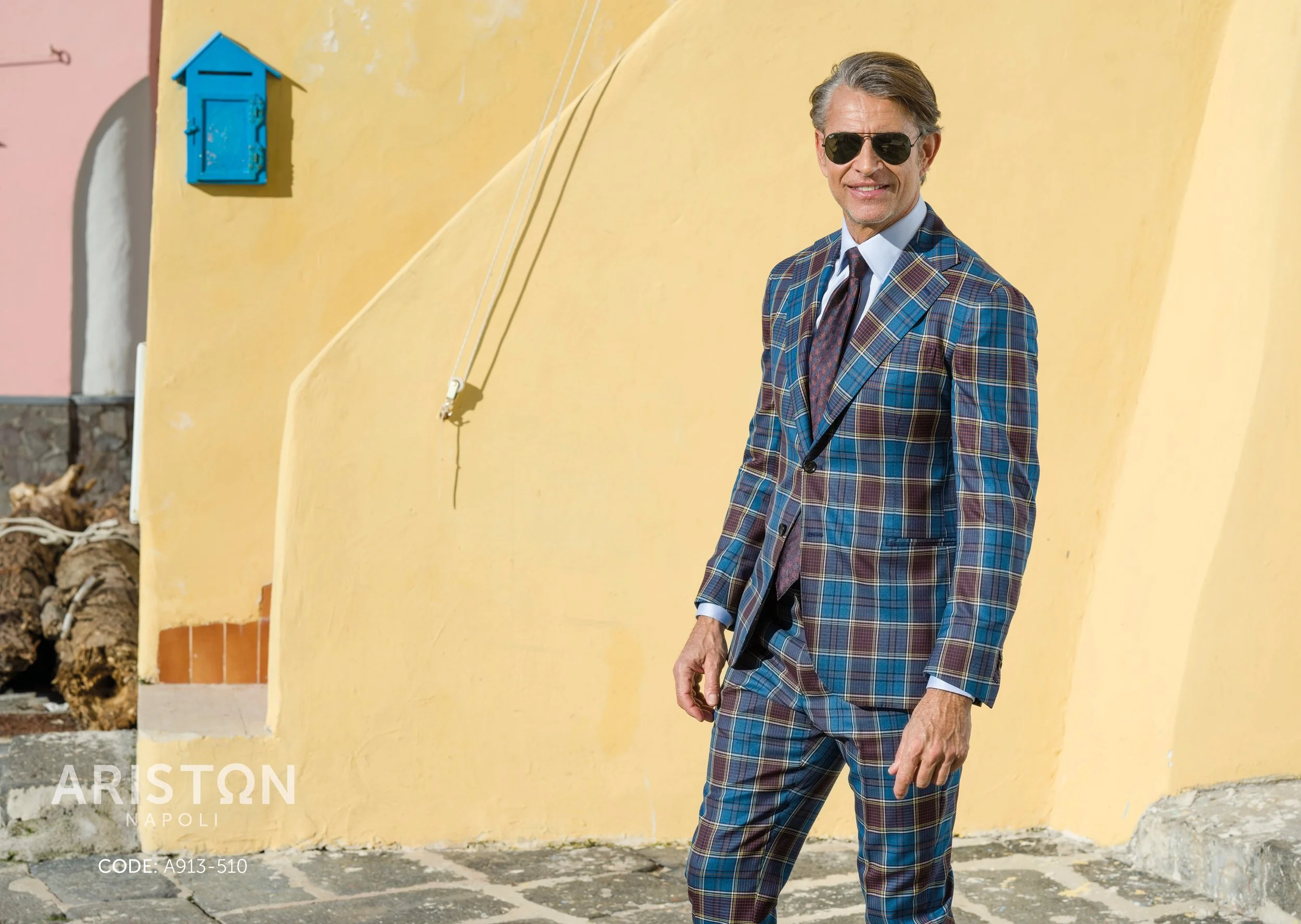 A man wearing a colorful plaid suit, sunglasses, and a tie, standing in front of a yellow wall with a blue mailbox decoration. The setting appears sunny and Mediterranean.