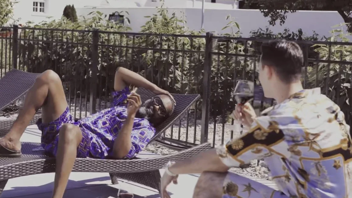 Two men enjoying relaxing outdoors on lounge chairs, one lying on a sunbed with sunglasses and colorful clothing, the other sitting with a glass of red wine, wearing a patterned shirt, sunny day.