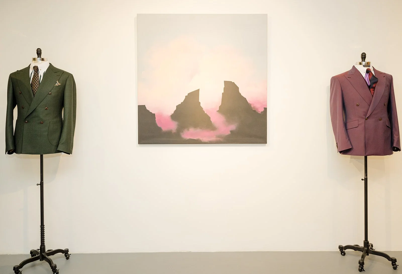 Two mannequins dressed in suits are displayed on either side of a framed painting with mountain peaks and pink and purple hues on a white gallery wall.