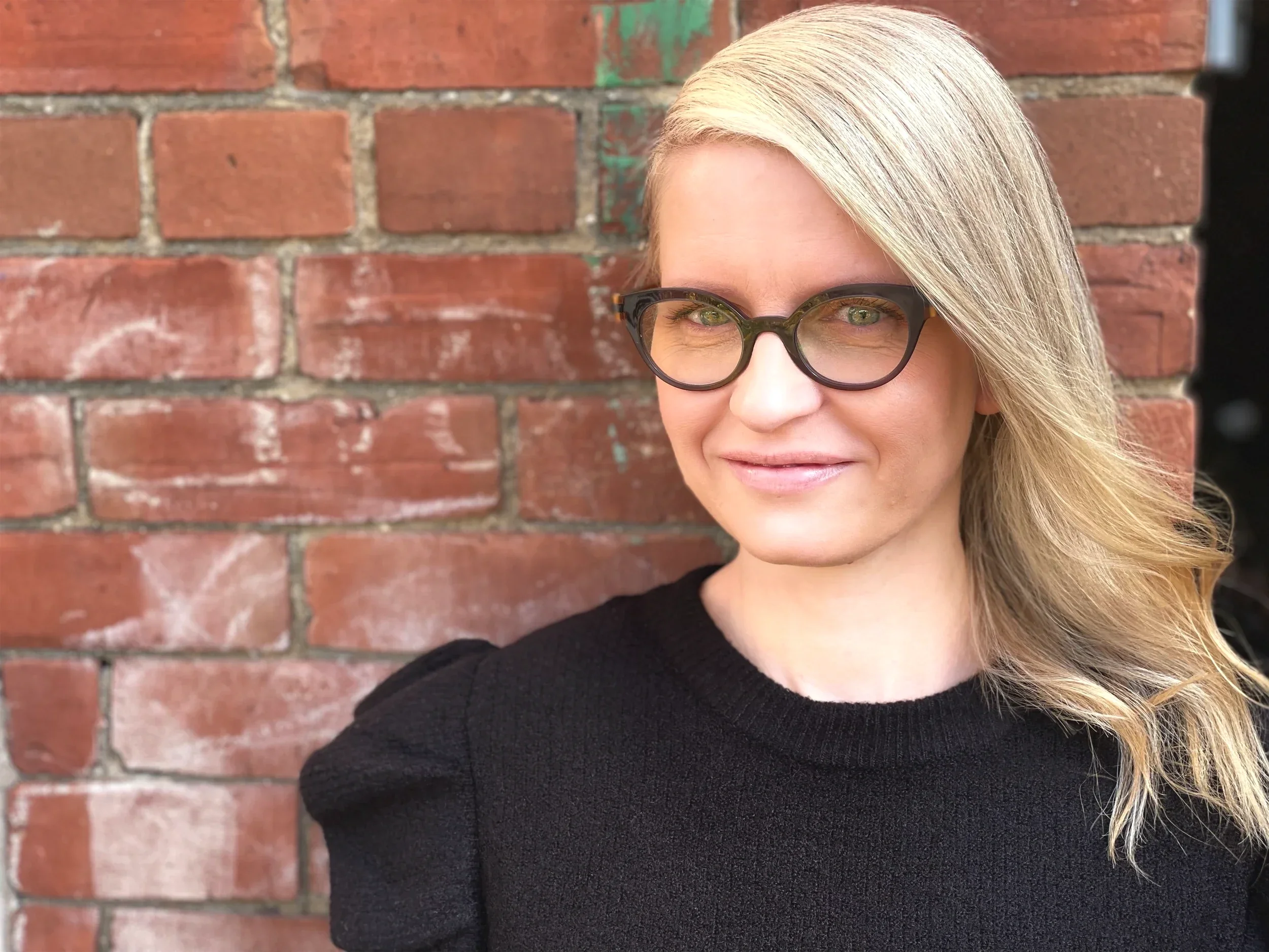 Close-up of a woman with blonde hair, glasses, and a black sweater standing in front of a red brick wall.