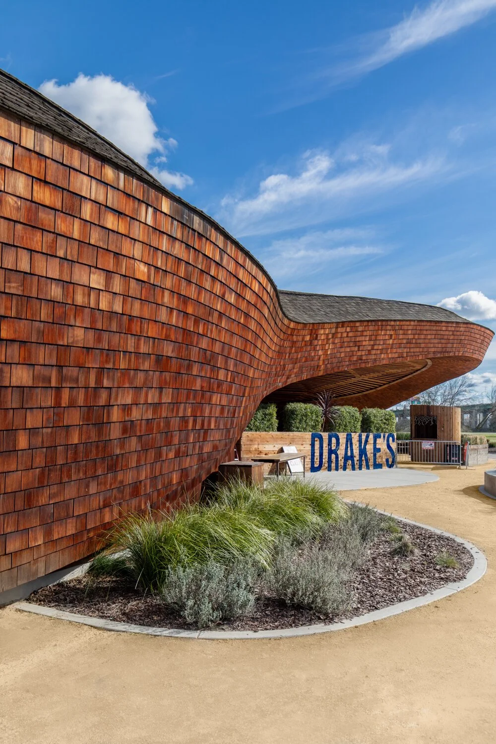 Exterior of Drakes: The Barn in Sacramento, California. A modern building with a curved wooden exterior, blue sky with scattered clouds in the background.