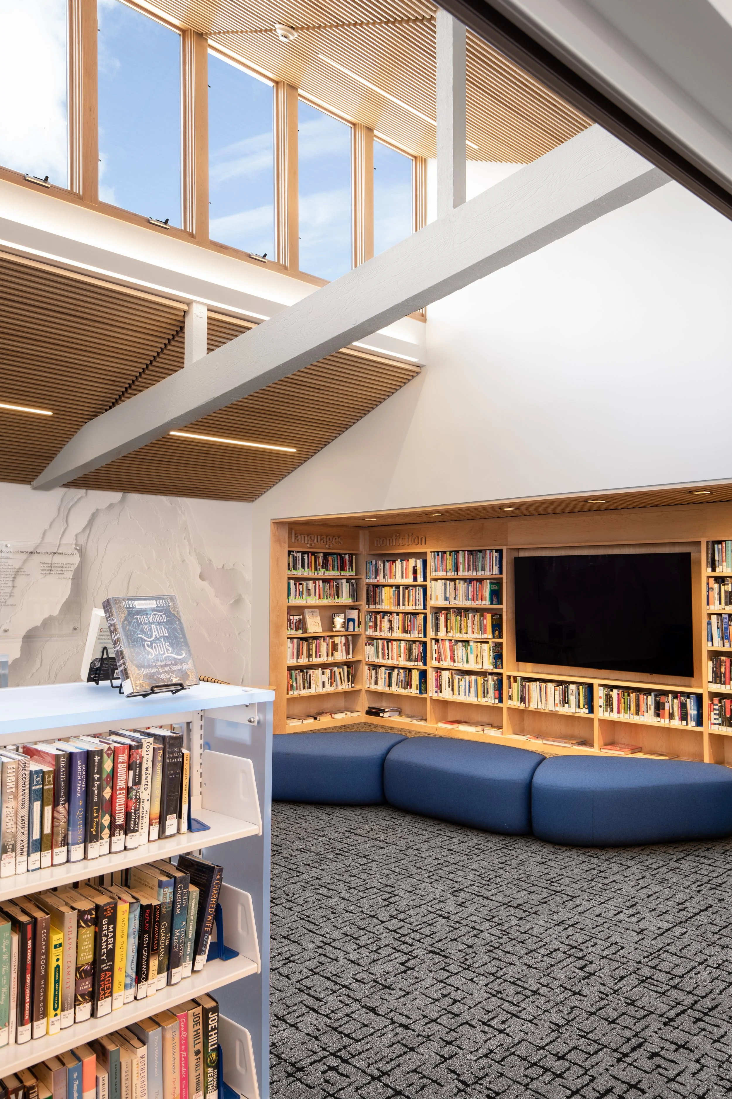 Interior of La Selva public library with a book display, built-in bookshelves filled with books, a large flat-screen TV, and a blue cushioned seating area. The ceiling features wooden slats and large windows allowing natural light.
