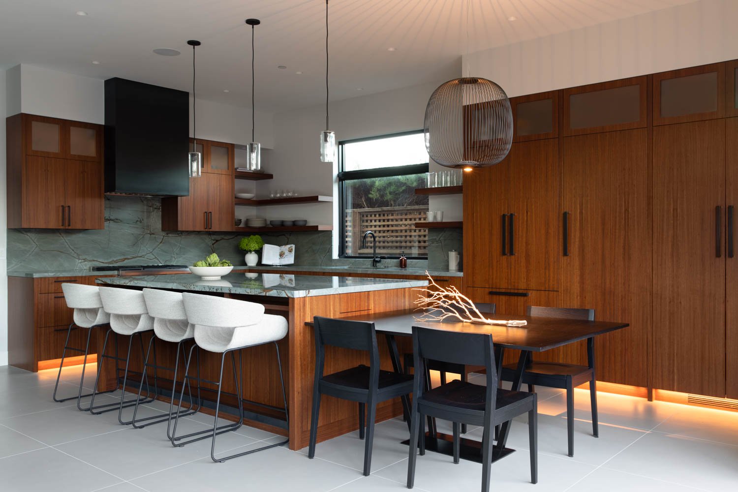 Modern kitchen with wooden cabinets, marble backsplash, island with white chairs, and a dining table with black chairs and a branch-shaped centerpiece.
