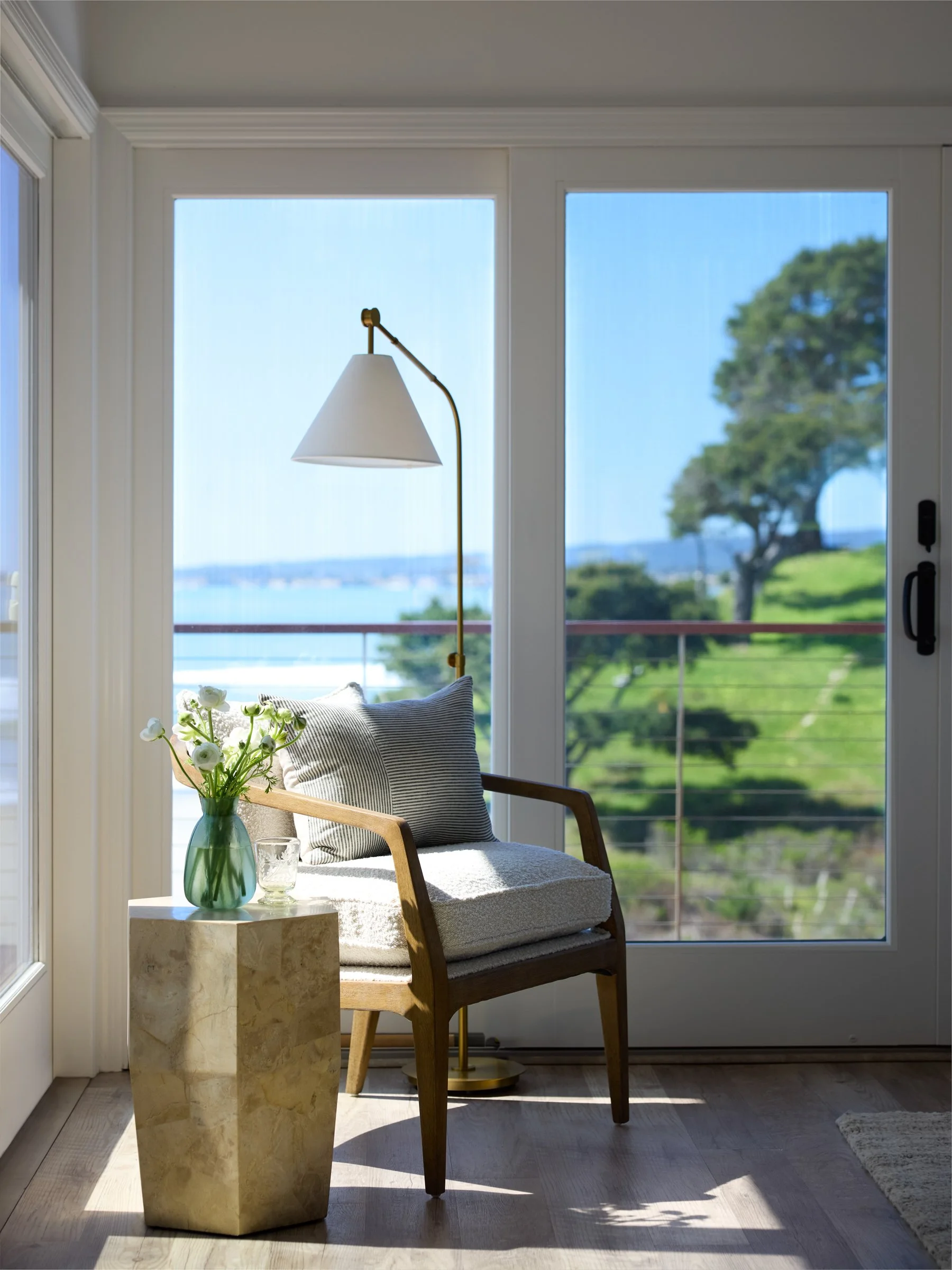 A cozy corner with a wooden armchair, a pillow, a glass vase with white flowers, a glass of water, a marble side table, and a brass standing lamp, overlooking a balcony with green landscape and a tree outside.