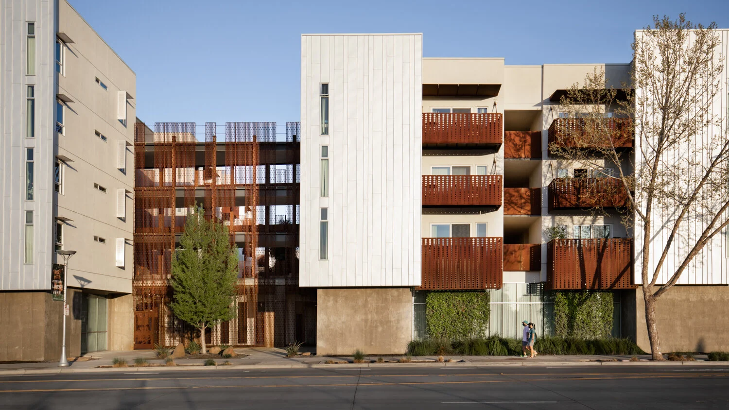 Modern multi-story apartment building with brown balconies, white panels, and with trees and two people walking on the sidewalk in front.