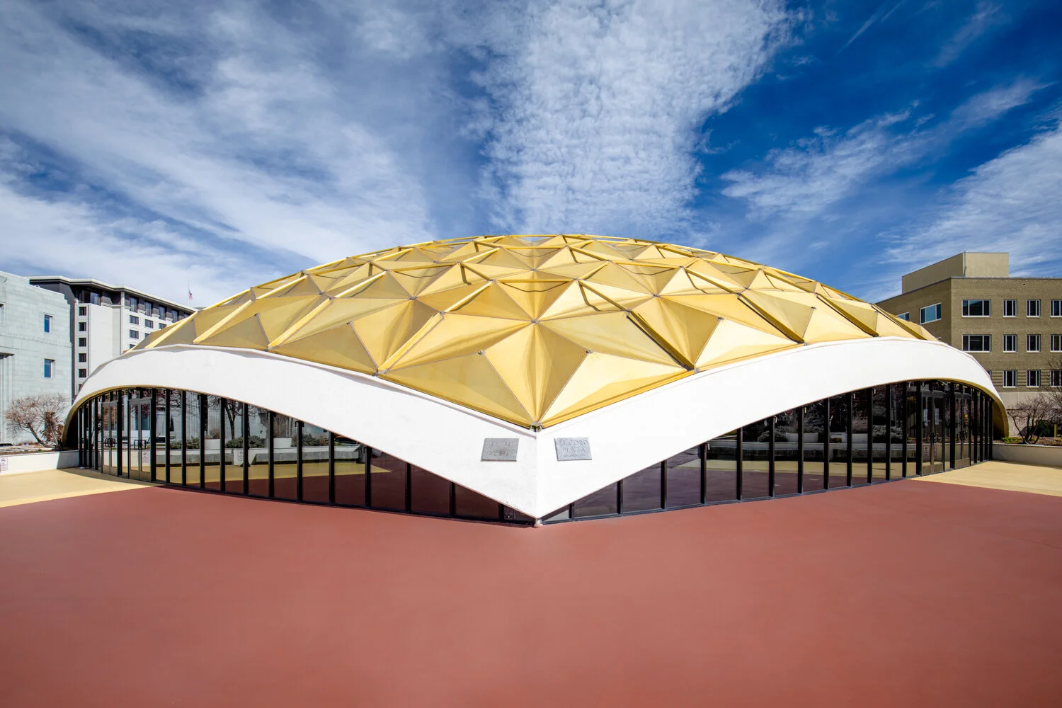 Pioneer center for Performing Arts in Reno, Nevada. A dome-shaped, yellow geometric roof, glass walls, and a red paved surrounding area, under a partly cloudy sky.