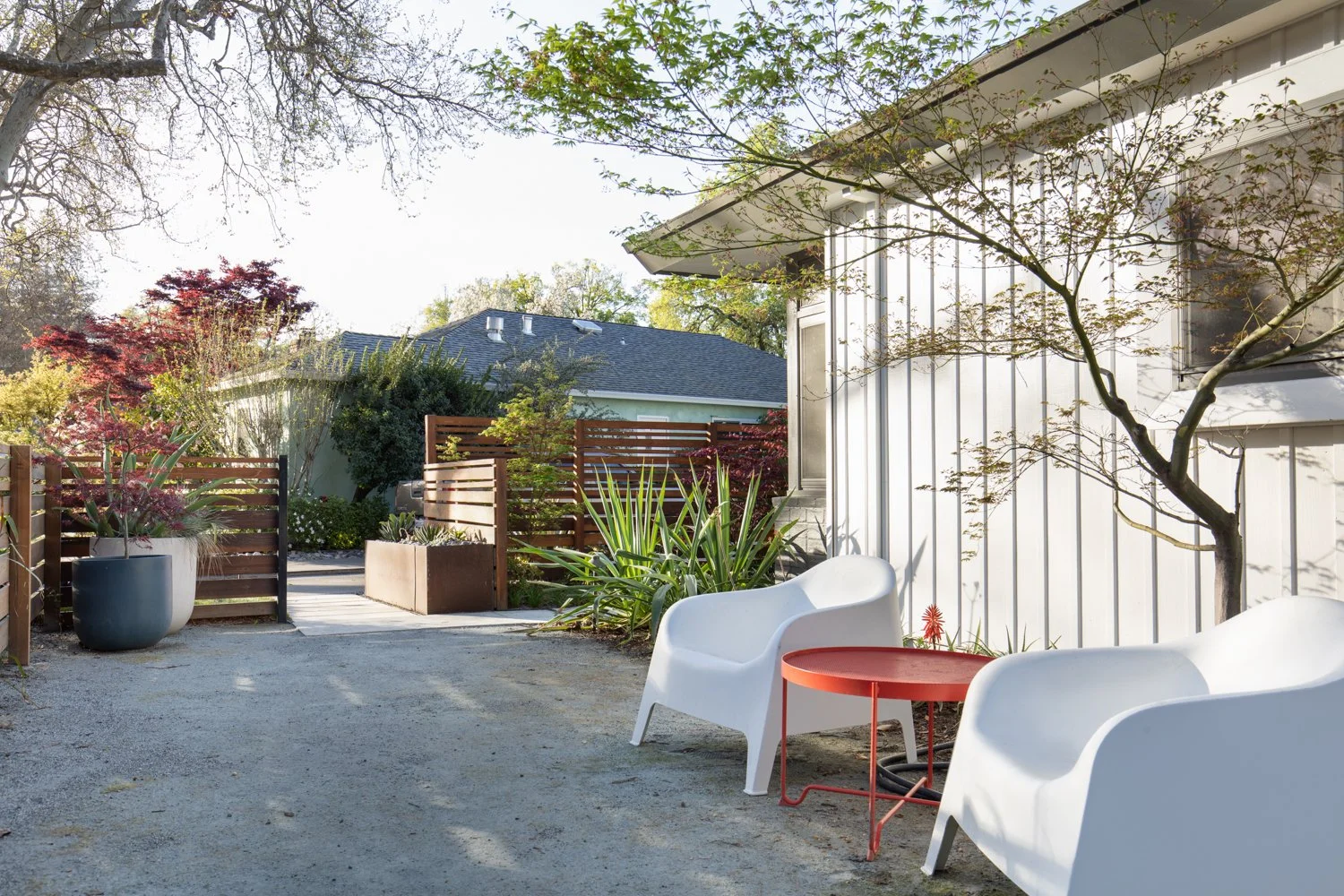 A modern backyard patio with two white outdoor chairs, a small red side table, and lush greenery including a small tree, potted plants, and a wooden fence, with a neighboring house in the background.