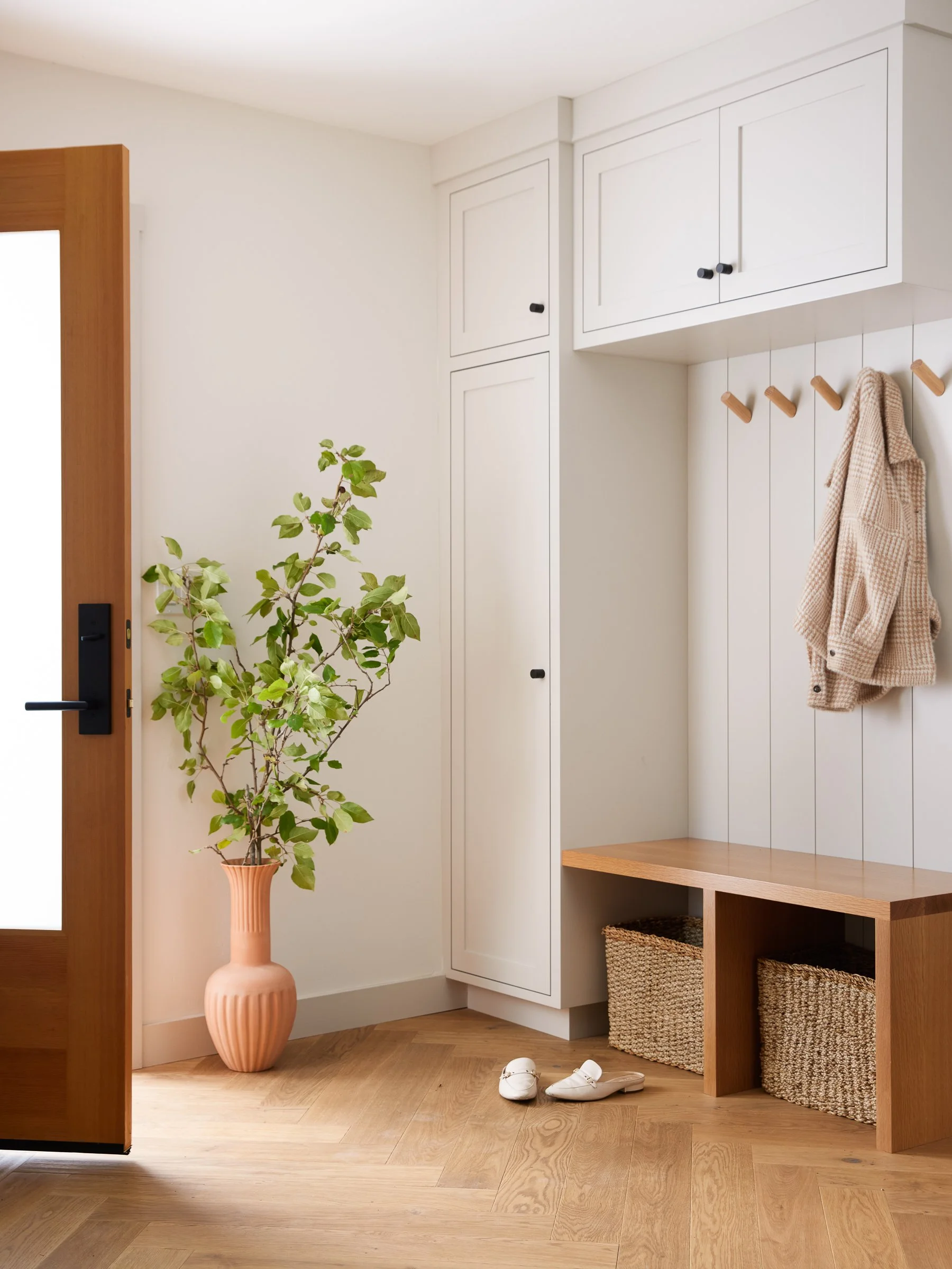 Entryway with a wooden door, white built-in cabinets, a pink vase with green plant, beige coat hanging on hooks, pair of white shoes on wooden floor, and storage baskets.